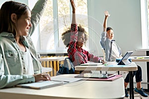 Diverse schoolkids sitting at desk raising hands answering at lesson.