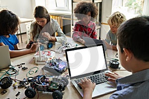 Diverse schoolkids at stem class building robot using pc with white screen.