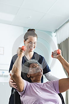 Diverse senior female patient exercising with weights and female doctor advising in hospital room