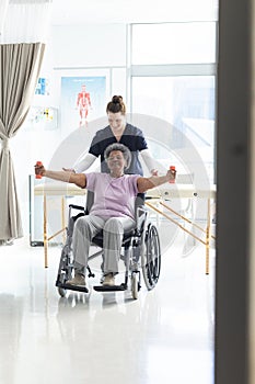 Diverse senior female patient exercising with weights and female doctor advising in hospital room