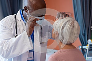 Diverse senior male doctor examining eye of senior female patient with penlight, copy space