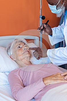 Diverse senior male doctor examining eye of senior female patient with penlight, copy space