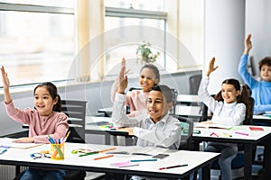 Diverse small schoolkids raising hands at classroom