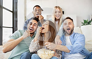 Diverse young friends eating popcorn while watching TV, having fun together at home