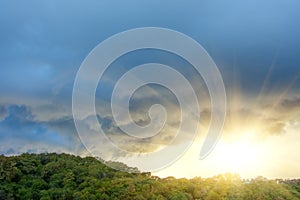 Divine rays from under dark storm clouds after a hurricane over a green forest hill with trees