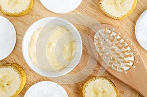 Diy banana mask face cream in the small white bowl, wooden hair brush, cotton pads and banana slices.