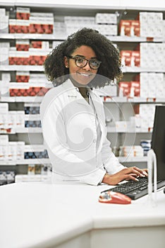 We do bookings as well. Portrait of a cheerful young female pharmacist typing on a computer while looking at the camera.