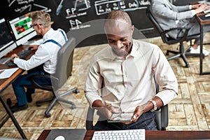 Do more, earn more. Cheerful african male trader sitting by desk and studying analytical reports using tablet pc in the
