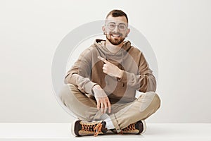 Do not miss chance to look left. Portrait of good-looking happy caucasian guy with beard and glasses sitting on floor