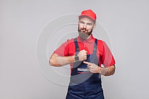We do ontime. Young confident handyman with beard in blue overall and red t-shirt standing and showing time on his wrist watch wi