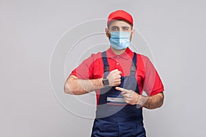 We do ontime. Young man with surgical medical mask in blue overall and red t-shirt standing and showing time on his wrist watch