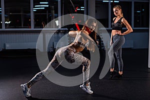 Do squats. Confident young personal trainer shows a slim athletic woman how to do squats on one leg with Trx fitness