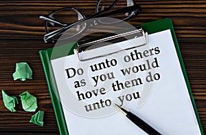 Do unto others as you would hove them do unto you - words on a white sheet on a brown wooden background with a pen and glasses