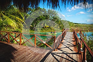 Dock along seven colored lagoon with canoe passing by surrounded by tropical plants in Bacalar, Quintana Roo, Mexico