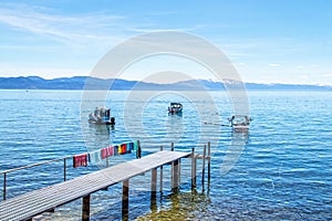 Dock with beach towels drying  stretching out onto Tahoe lake with boats moored offshore and snowtopped mountains and a pastel sky