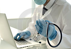 Doctor in blue gloves holds a stethoscope in one hand with the other hand is typing on a laptop in a hospital room