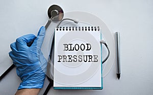 A doctor in blue medical gloves holds a notebook with the inscription BLOOD PRESSURE, on the background of a light table, along