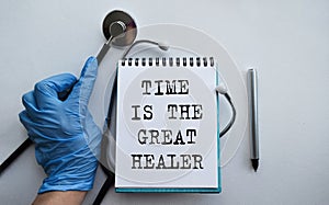 A doctor in blue medical gloves holds a notebook with the inscription TIME IS THE GREAT HEALER, on the background of a light table