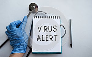 A doctor in blue medical gloves holds a notebook with the inscription VIRUS ALERT, on the background of a light table, along with