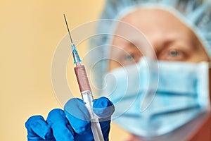 A doctor in blue medical gloves, medical mask and safety cap is holding a syringe. Closeup