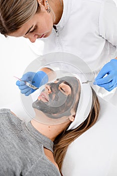 Doctor in a blue rubber gloves makes a face mask for a woman with the help of cosmetics tools