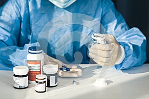 Doctor in a blue surgical gown and mask holds in his hand a medicine bottle. Many pills and drugs on white table, healthcare