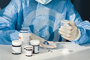 Doctor in a blue surgical gown and mask holds in his hand a medicine bottle. Many pills and drugs on white table, healthcare