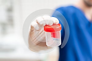 Doctor in blue uniform and latex gloves is holding an empty plastic container for taking urine samples, light background