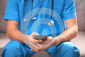 A doctor in a blue uniform with a stethoscope around his neck is sitting on a sofa with a phone in his hands