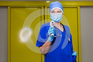 A doctor in a blue uniform, surgical cap, mask and gloves holding a non-contact thermometer