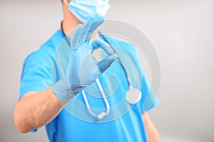 A doctor in a blue uniform wearing a medical mask with a stethoscope around his neck shows the Ok gesture  against a uniform gray