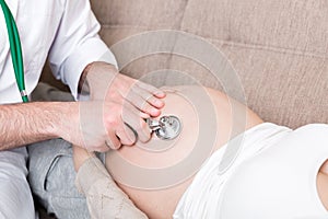 Doctor is checking pregnant woman`s belly with a stethoscope. Physician is listening to a heartbeat of a baby