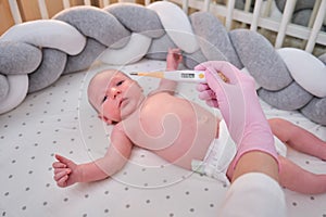 Doctor checks the temperature of the newborn baby with a thermometer. A nurse in uniform measures the child fever with a