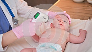 Doctor checks the temperature of the newborn baby with a thermometer. A nurse in uniform measures the child fever with a