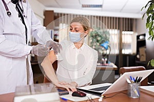 Doctor in coat and protective mask giving vaccine injection to woman manager