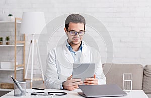Doctor consultation online. Young man in glasses and white coat with wireless headphones looks at tablet