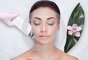 The doctor-cosmetologist makes the ultrasound cleaning procedure of the facial skin of a beautiful, young woman in a beauty salon.