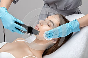 The doctor-cosmetologist makes the ultrasound cleaning procedure of the facial skin of a beautiful, young woman in a beauty salon