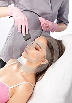 The doctor-cosmetologist makes the ultrasound cleaning procedure of the facial skin of a beautiful, young woman in a beauty salon