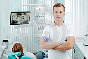 Doctor dentist smiling and standing with hands crossed in dental clinic on backdrop with medical equipment, x-ray dental and