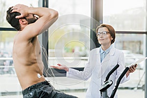 doctor with digital tablet gesturing near shirtless sportsman during endurance test in gym.