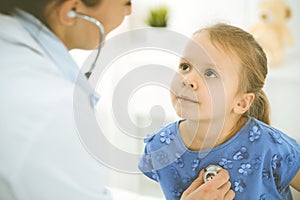 Doctor examining a child by stethoscope. Happy smiling girl patient dressed in blue dress is at usual medical inspection