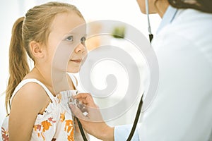 Doctor examining a child by stethoscope. Happy smiling girl patient dressed in bright color dress is at usual medical
