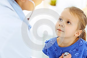 Doctor examining a child by stethoscope in sunny clinic. Happy smiling girl patient dressed in blue dress is at usual