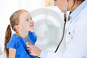 Doctor examining a child by stethoscope in sunny clinic. Happy smiling girl patient dressed in blue dress is at usual