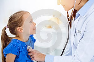 Doctor examining a child by stethoscope in sunny clinic. Happy smiling girl patient dressed in blue dress is at usual