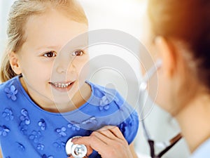 Doctor examining a child by stethoscope in sunny clinic. Happy smiling girl patient dressed in blue dress is at usual