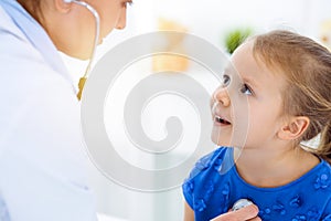 Doctor examining a child by stethoscope in sunny clinic. Happy smiling girl patient dressed in blue dress is at usual