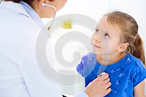 Doctor examining a child by stethoscope in sunny clinic. Happy smiling girl patient dressed in blue dress is at usual