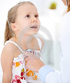 Doctor examining a child by stethoscope in sunny clinic. Happy smiling girl patient dressed in bright color dress is at